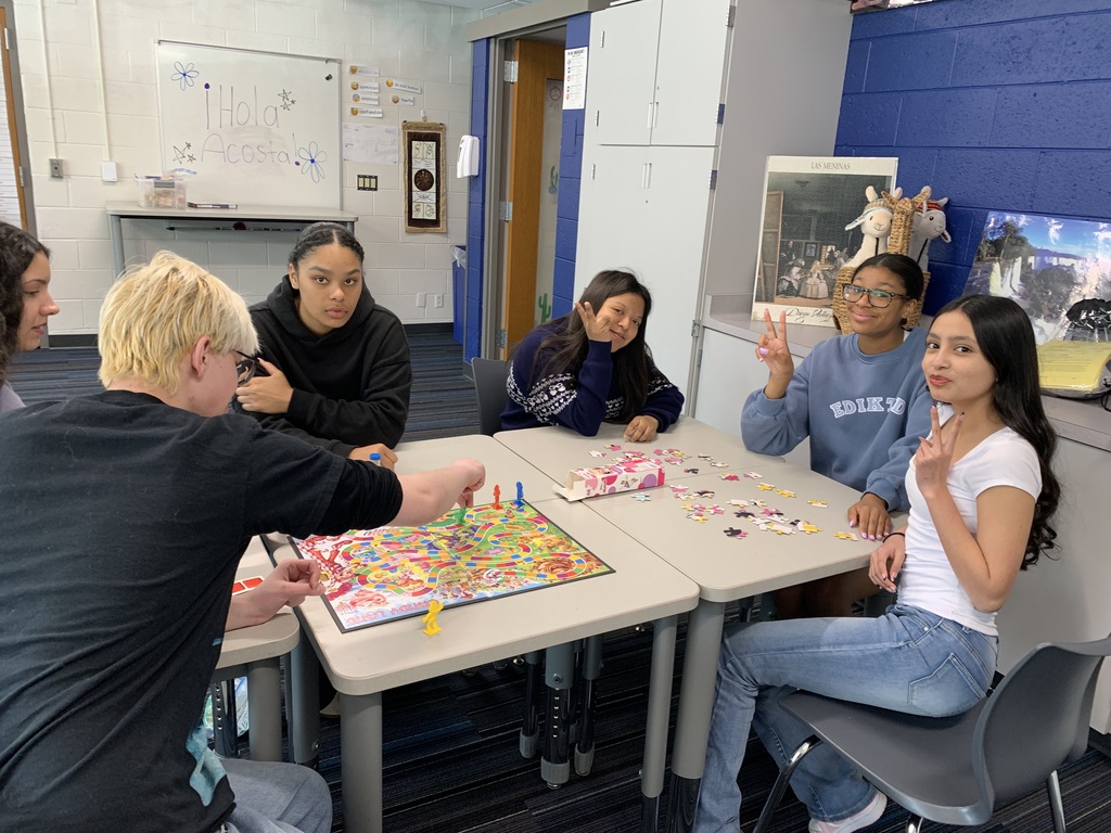 A group of people playing a board game in a classroom. A whiteboard is visible on the wall.