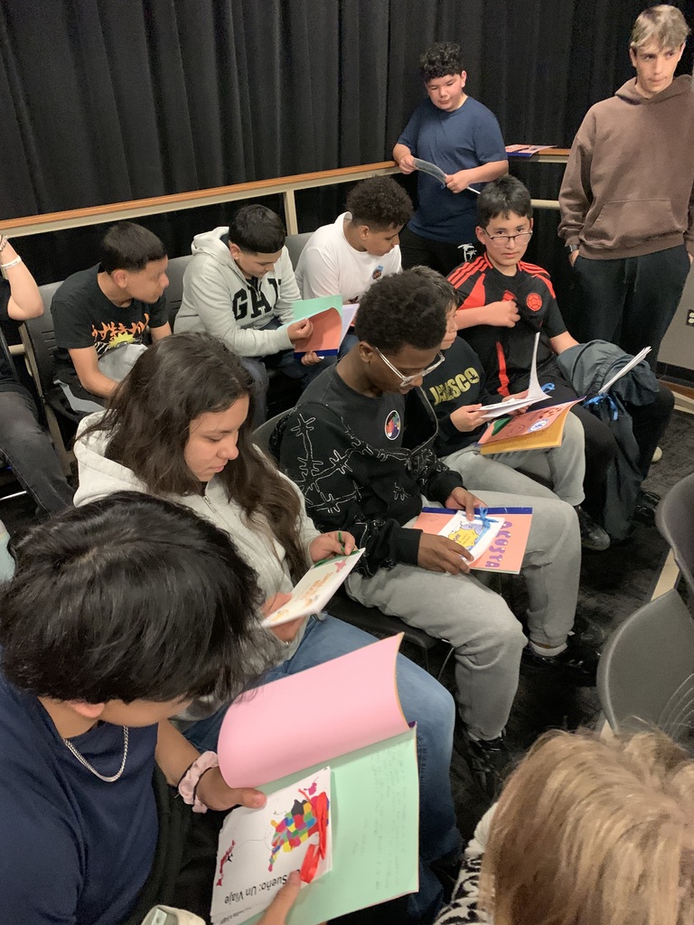 A group of individuals in a classroom setting, seated on chairs and holding books, with some reading.