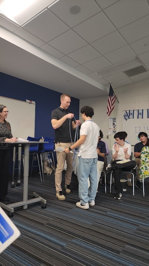 A classroom with blue walls and white ceiling. A group of people are gathered, with one person demonstrating to others.