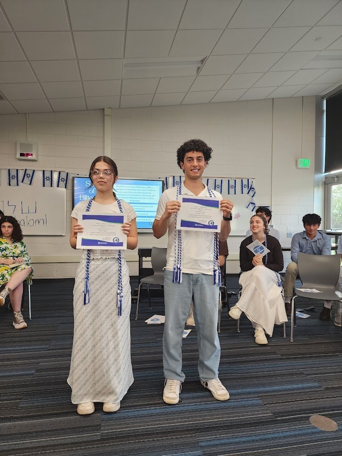 Two people stand in a room holding certificates; one wears glasses and a white dress, the other in pants and a white shirt.