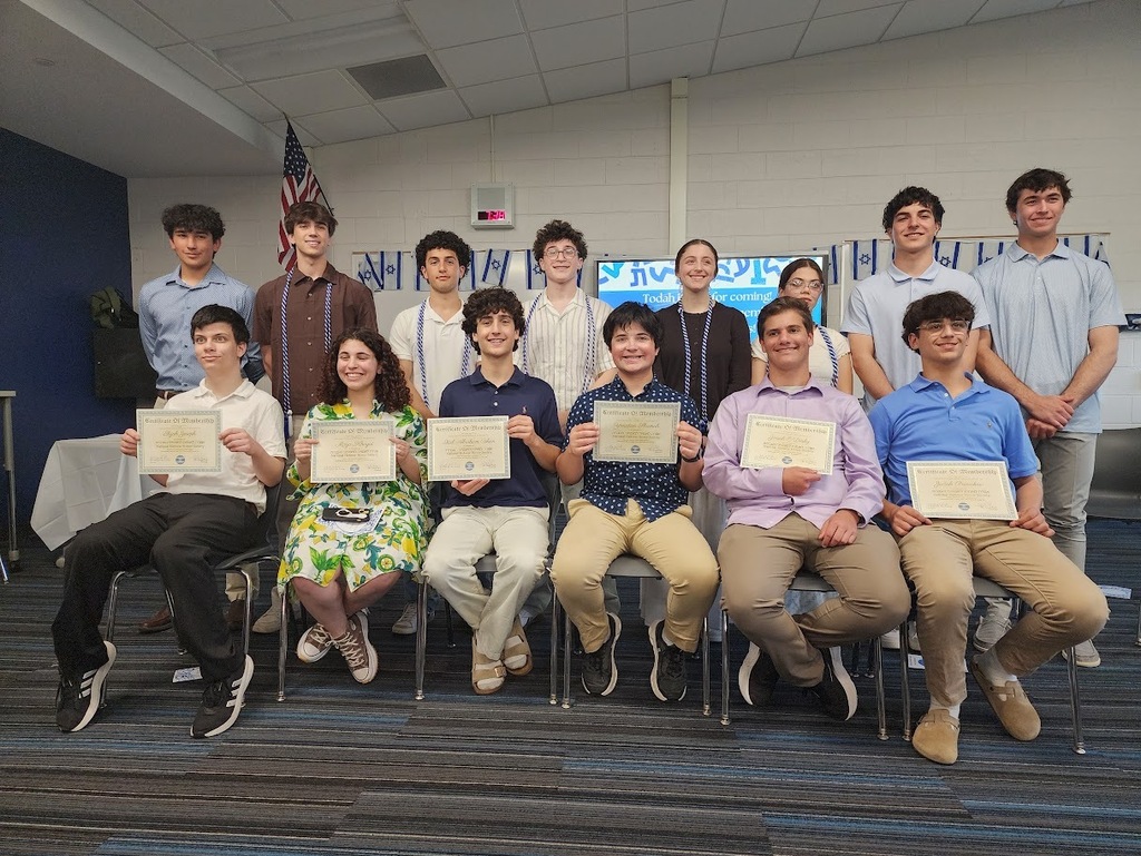 A group of students seated in chairs, holding certificates, posing for a photo in a room with a flag and table.