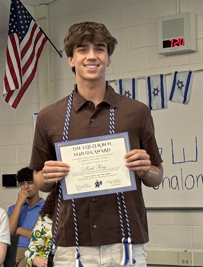 A man wearing a brown shirt and white pants holds a certificate with tassels, smiling. A flag hangs on the wall behind him.