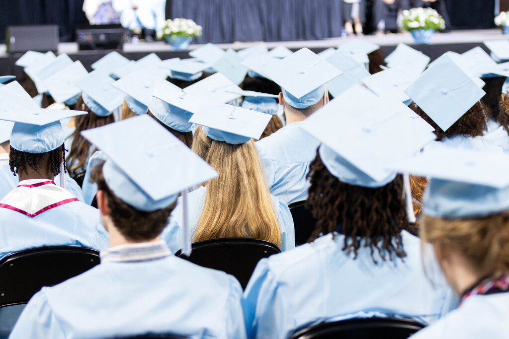 Graduates in caps and gowns sit in rows at an event, facing the stage. The stage has blue curtains and potted plants.