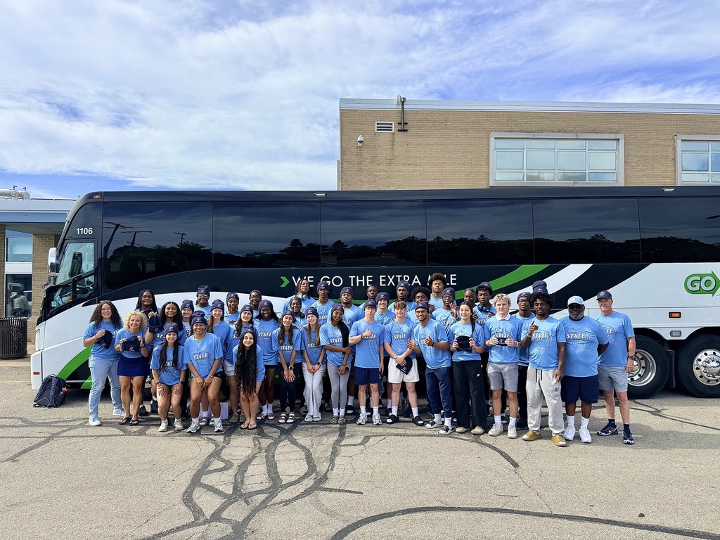 Group of young people in blue shirts, standing in front of a bus with a white and green design.