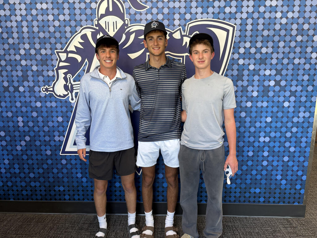 Three individuals stand together against a blue background with a logo. Two wear light blue and gray shirts, and the third wears a darker gray polo.