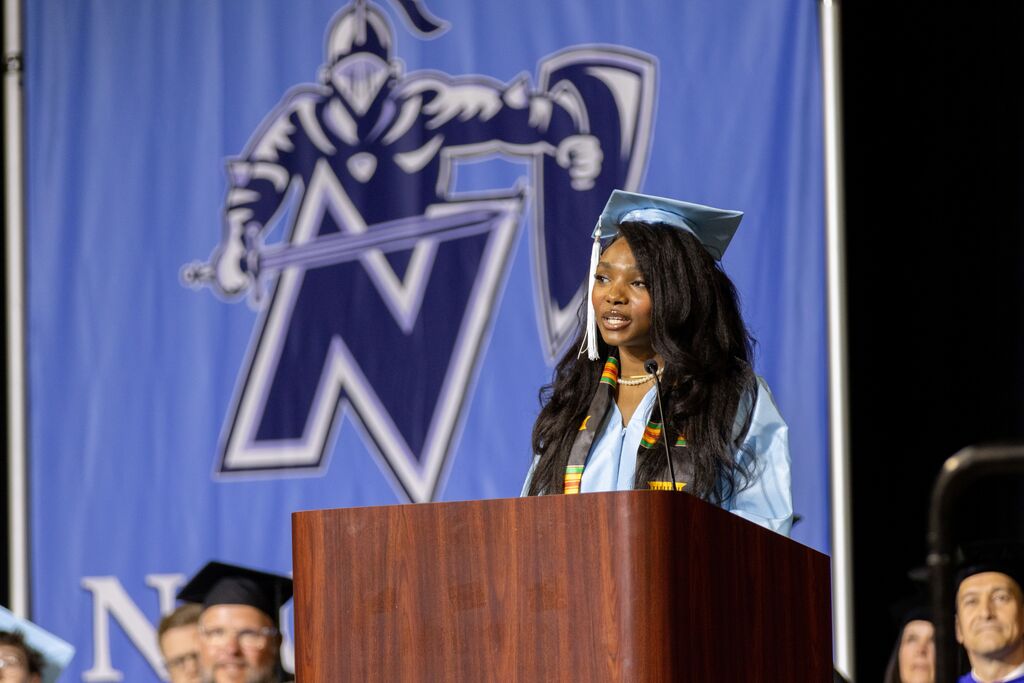A woman in a cap and gown gives a speech at a podium. Behind her is a banner with a logo.