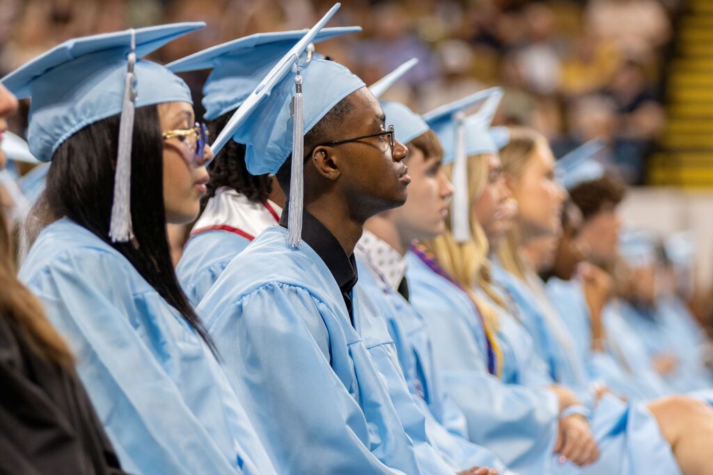 A group of people in blue graduation gowns and caps sitting in an audience, some looking forward.