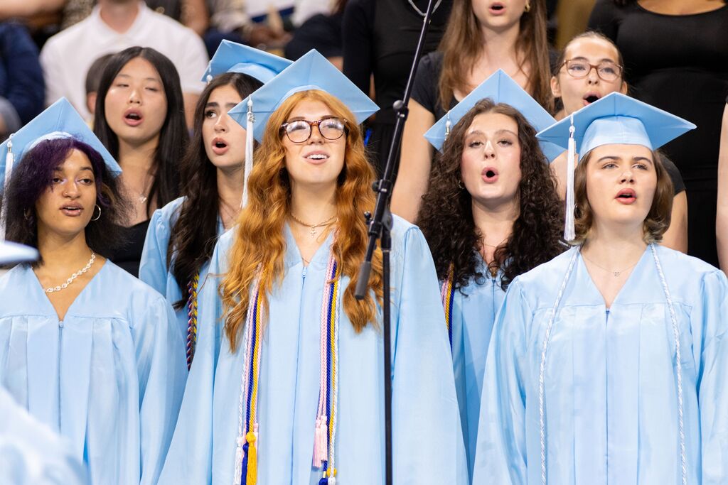 A choir of graduates in blue gowns and caps sings in an auditorium, with an audience behind them.