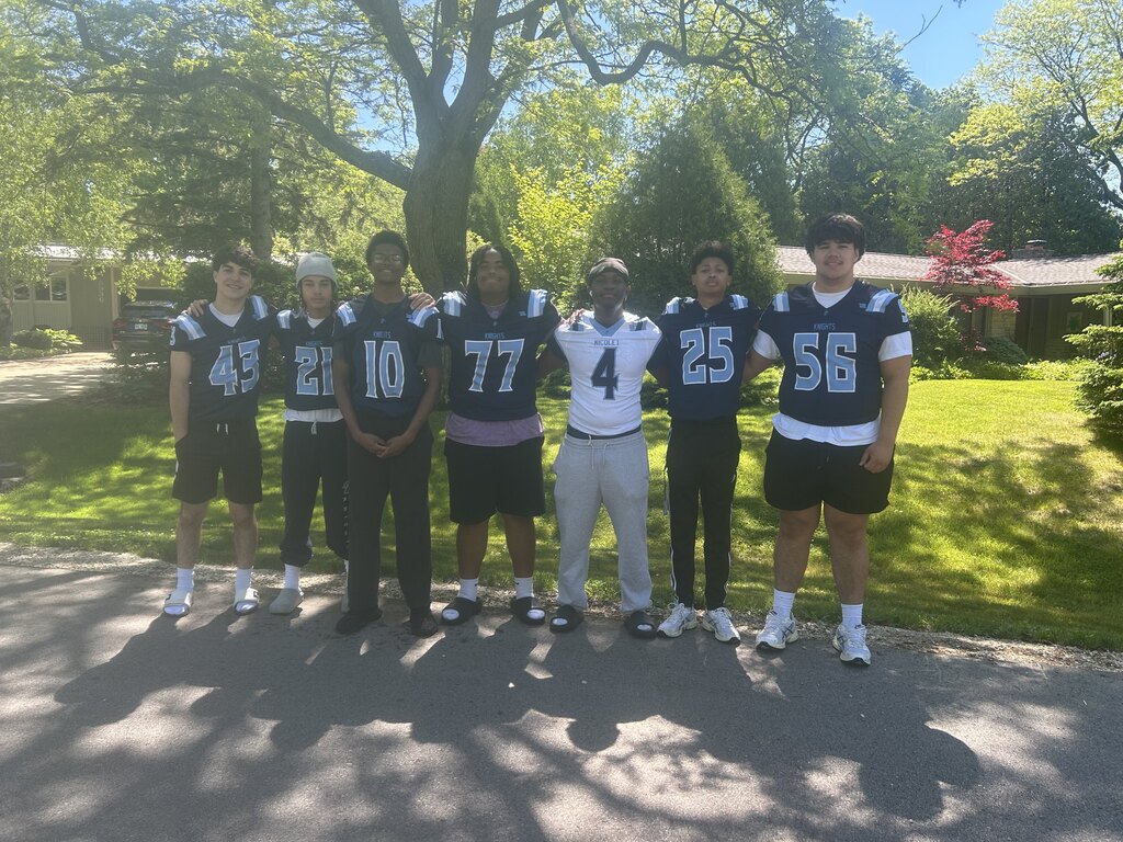 Group of six athletes in jerseys numbered 43, 21, 10, 77, 4, 25, and 56 stand on a driveway.