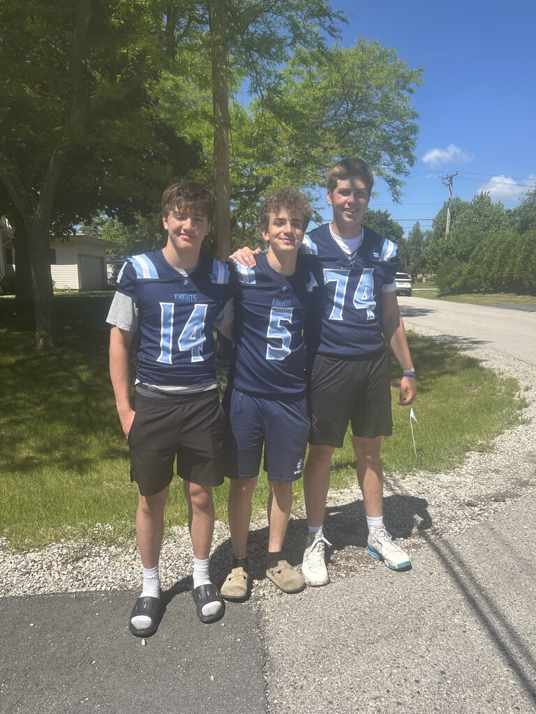 Three boys in blue jerseys with numbers 14, 5, and 74 pose together on a road.