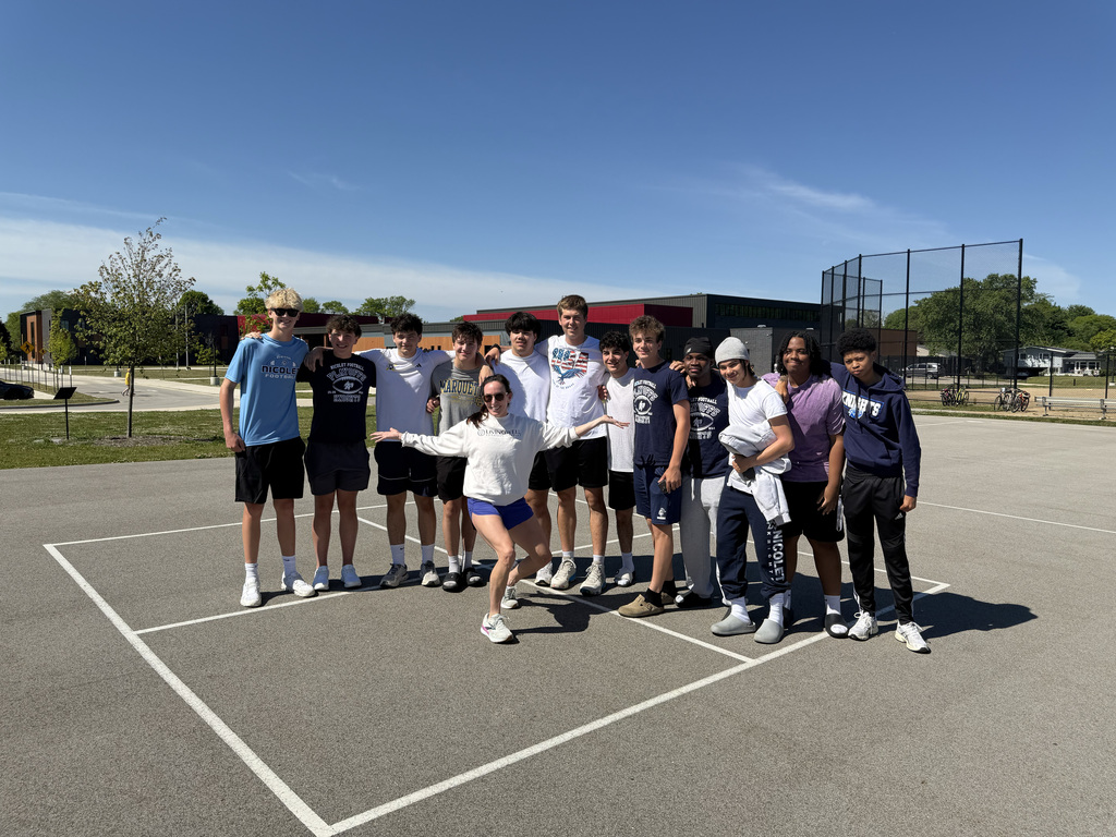 A group of people stand on a tennis court, wearing various athletic clothing. They pose for a photo in clear weather.