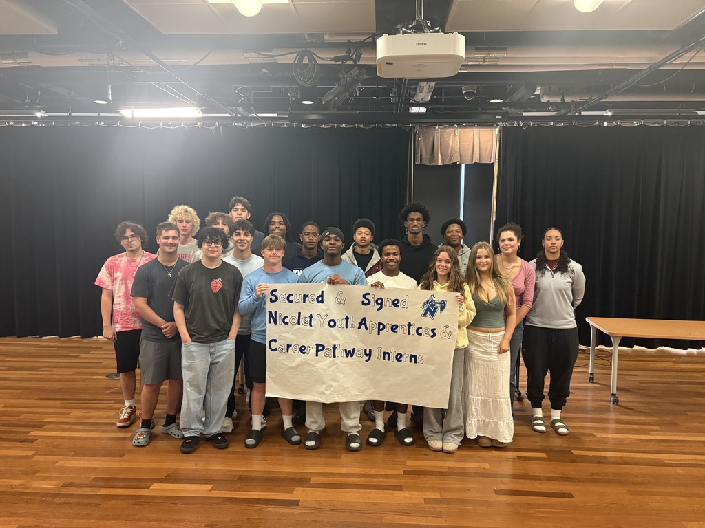 A group of people stand in a room with a banner, wooden floor, and black curtains.