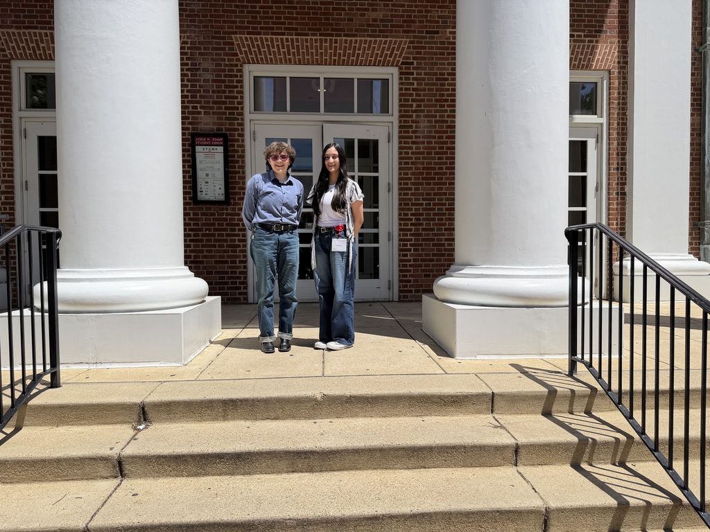 Two people stand in front of a brick building with white pillars and a staircase. One wears a blue shirt and jeans, the other a white top and jeans.