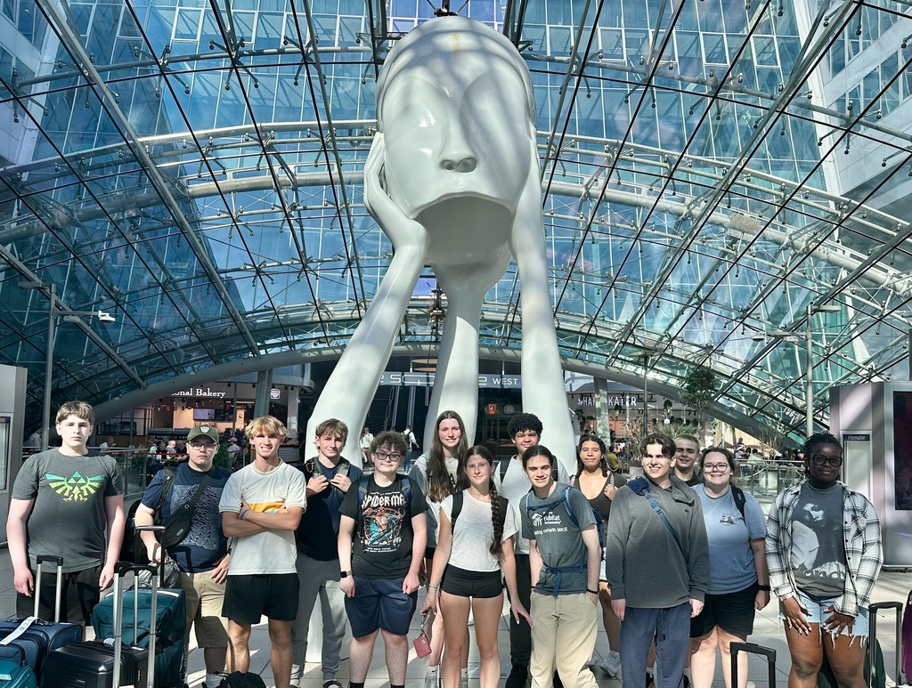 A group of people stands before a large white sculpture in a modern, glass-roofed atrium.