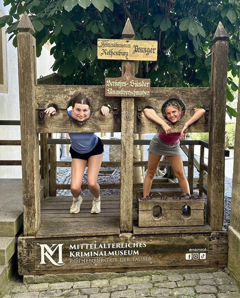 Two young girls pose with arms in a wooden pillory outside a museum, green foliage behind.