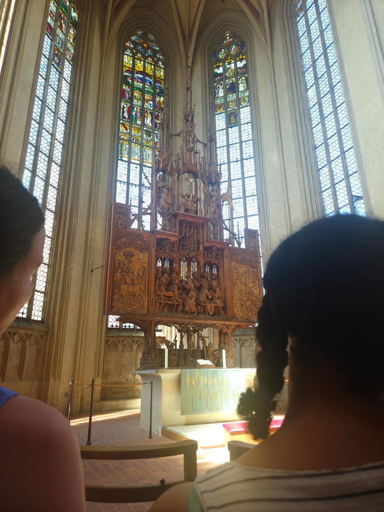 Two people gaze at a wooden altarpiece with intricate carvings and stained glass windows behind them.