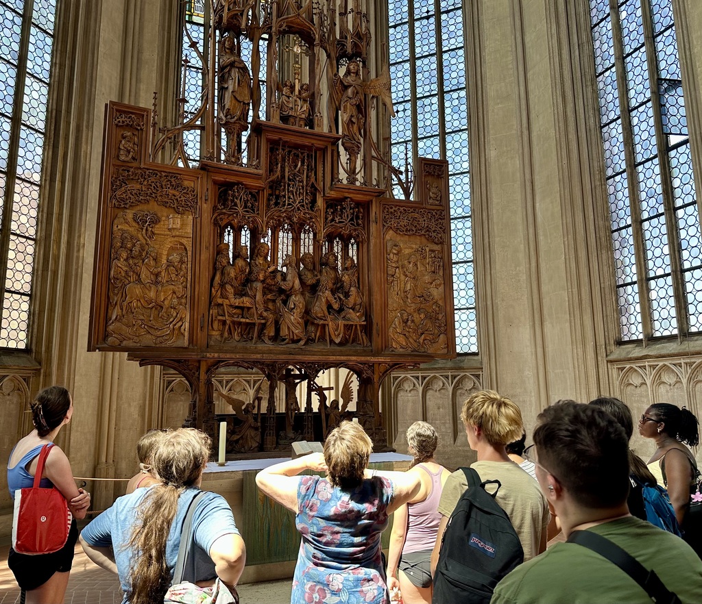 Visitors in a cathedral with a large altarpiece behind them. The altarpiece is ornate and depicts various scenes.