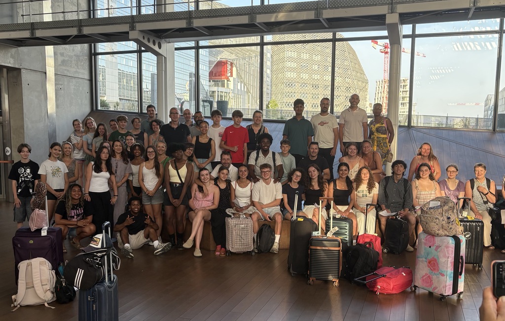 A group of people posing for a picture with luggage and suitcases in a room with large windows.