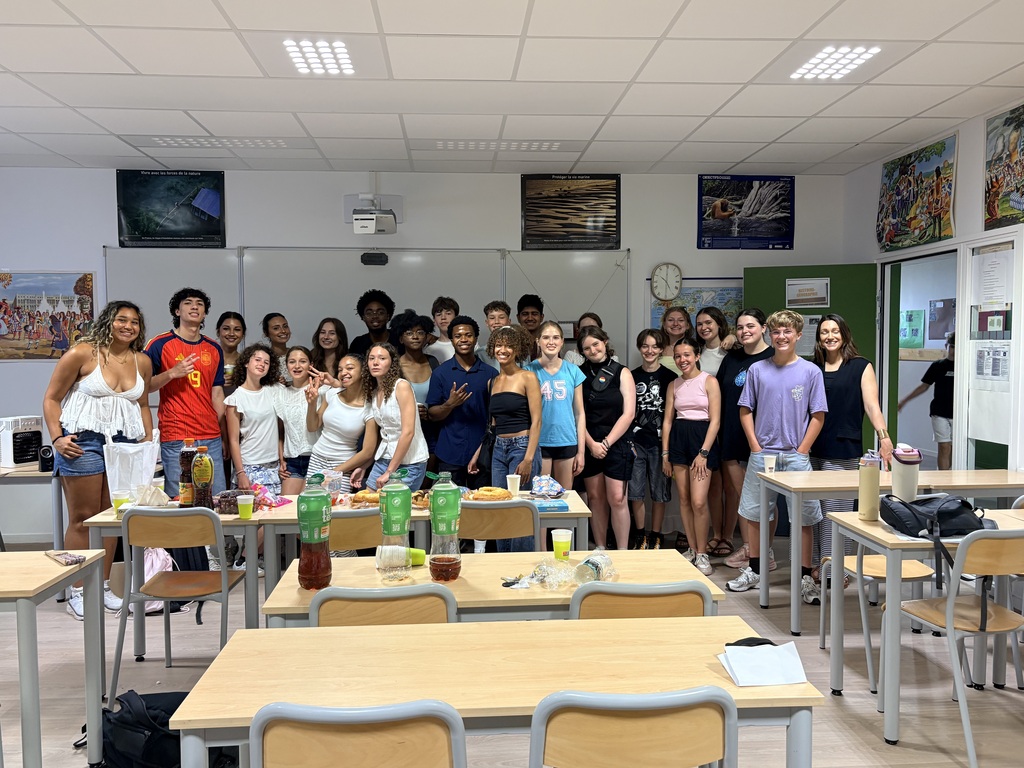 A classroom with tables, chairs, and people. Some have bottles on tables. Artwork hangs on the walls.