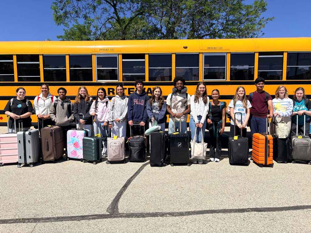 A group of students pose for a photo in front of a yellow school bus. They are all holding suitcases.