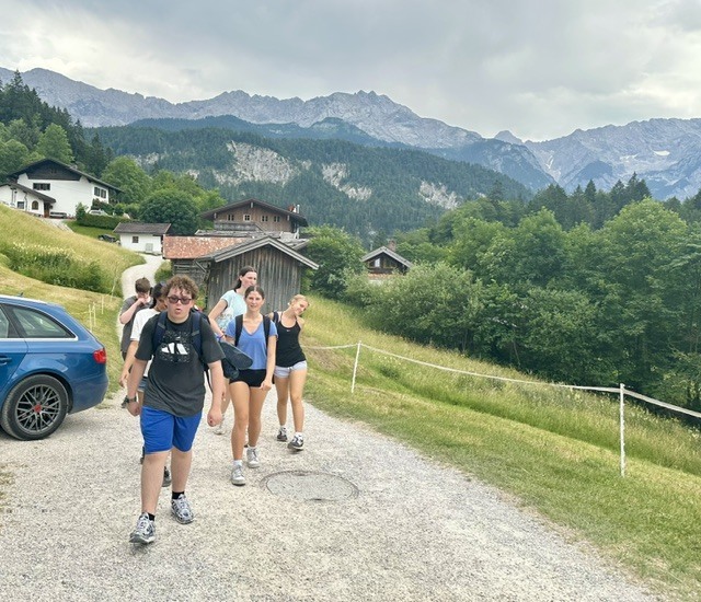 A group of people hiking on a path, with a blue car parked, houses, and mountains in the background.