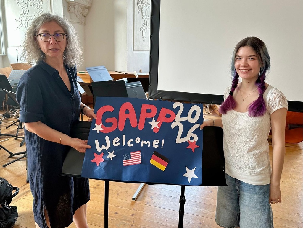Two women hold a welcome sign with USA and Germany flags in a room with musical instruments.
