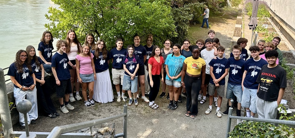 A group of young people stands on concrete next to a river, wearing similar blue shirts and casual outfits.
