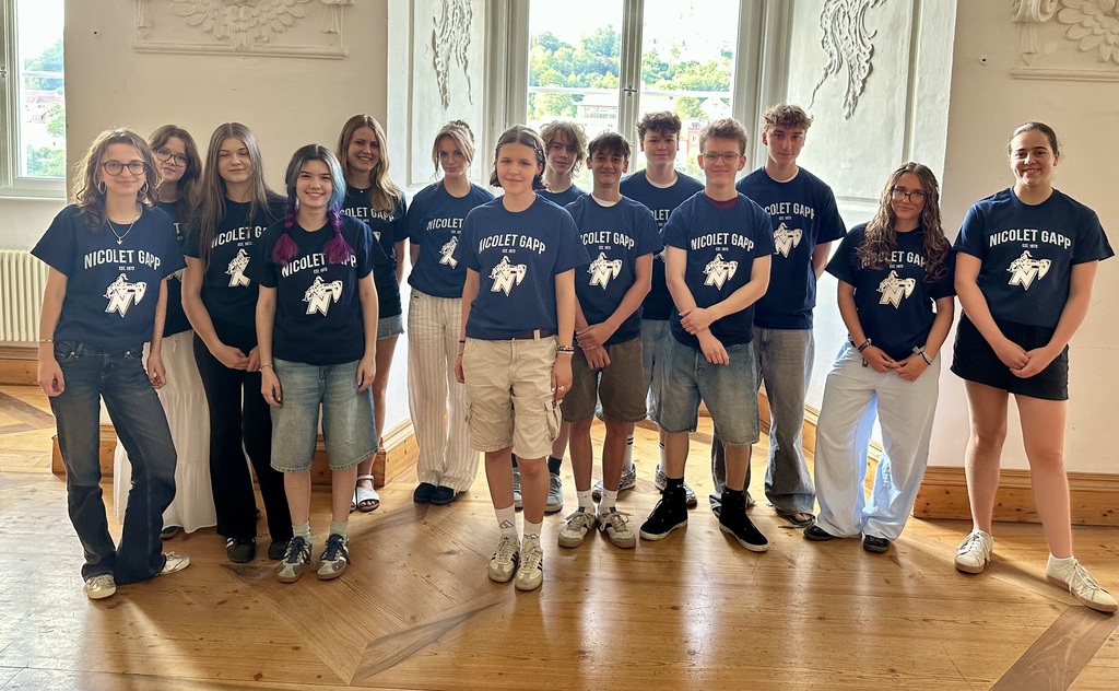 A group of young people in matching shirts, standing in a room with wooden floors and large windows.