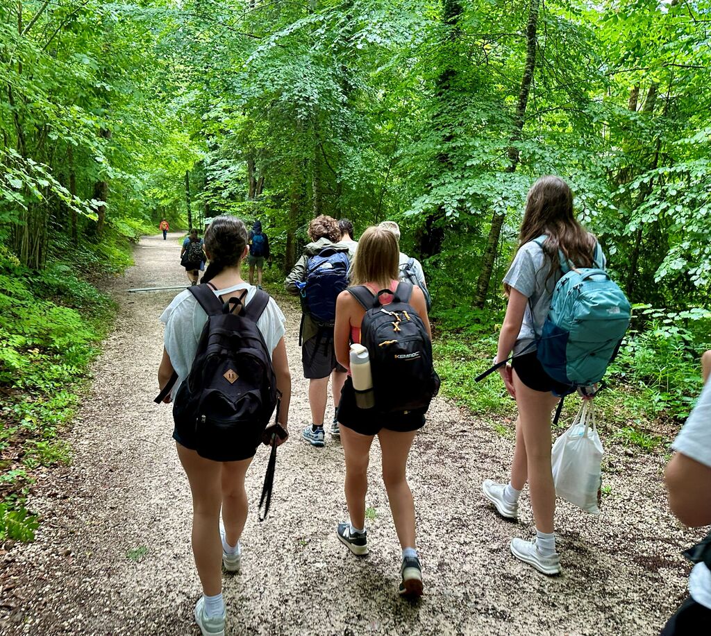 Five people walk along a dirt path in a forest, carrying backpacks. Some hold plastic bags.
