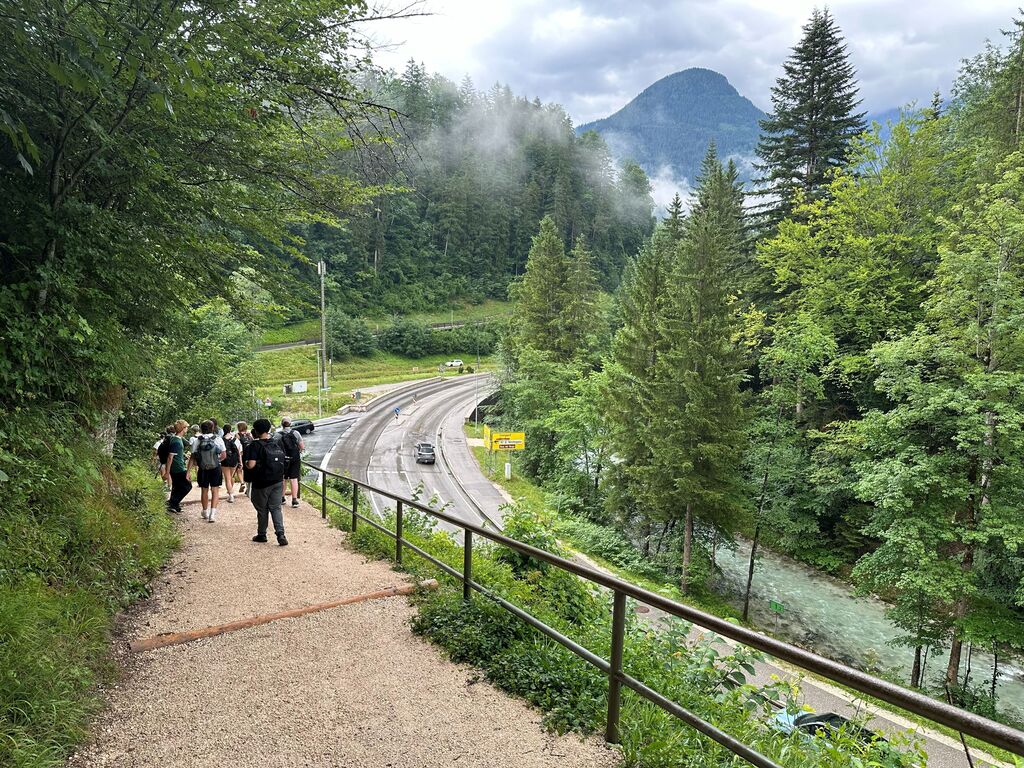 A group of people walk on a path next to a guardrail. A road and river are visible. Trees surround the area, and mountains are in the distance.