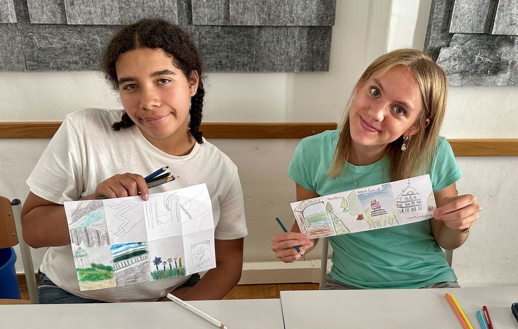 Two girls display artwork. One holds a drawing with cityscape, other holds a drawing with trees.