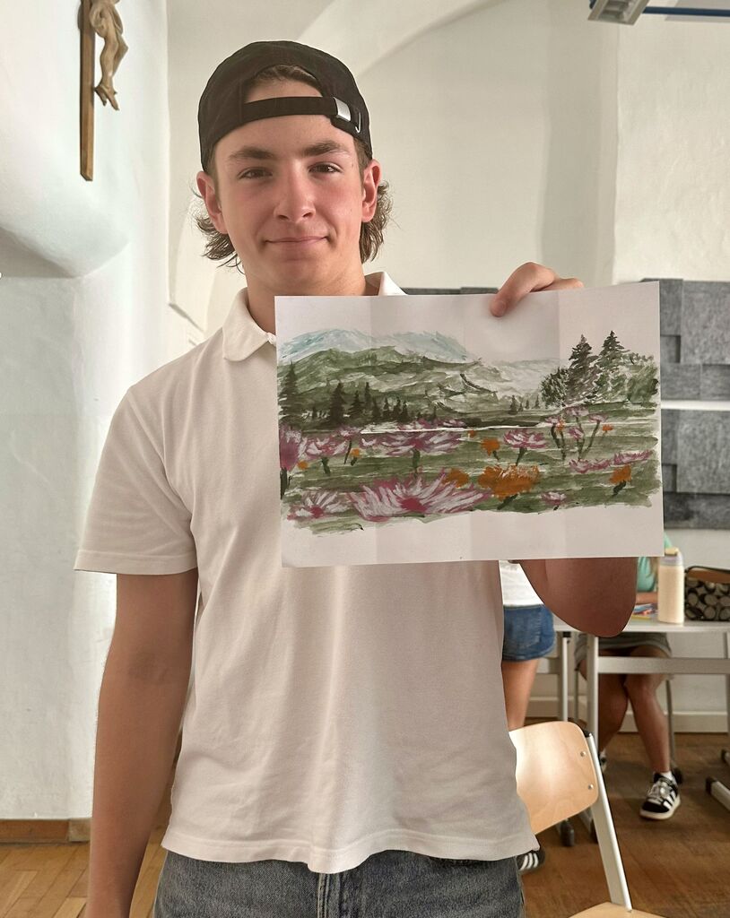 Man with a cap holds a small watercolor painting of a landscape, standing in a room.