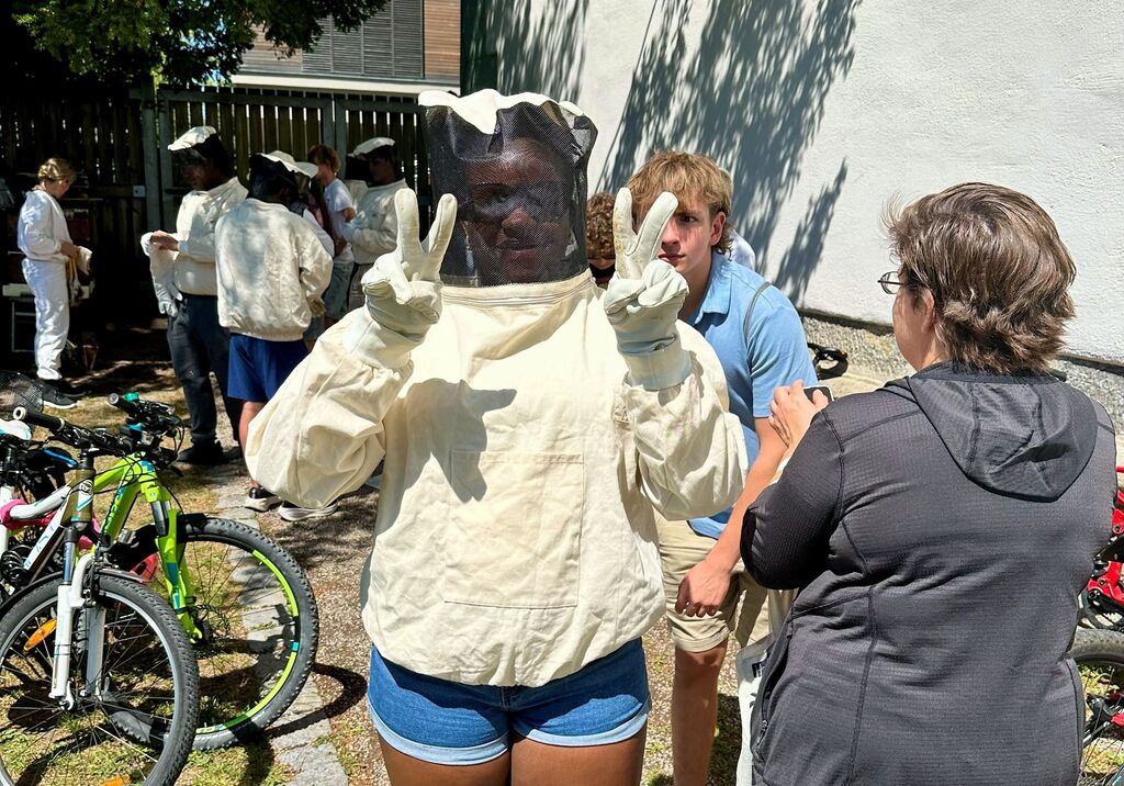 People in protective beekeeping gear outside. A woman in front wears a mask and glove. Behind her, others stand near bicycles.