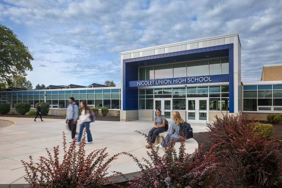 Exterior of a modern school building with blue accents. Several individuals are walking or sitting outside.