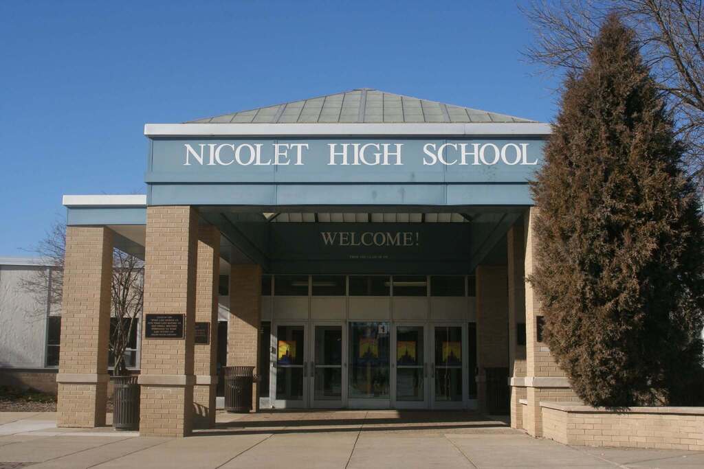 Entrance to Nicolet High School with a blue sign, glass doors, and a tree in front under a clear sky.