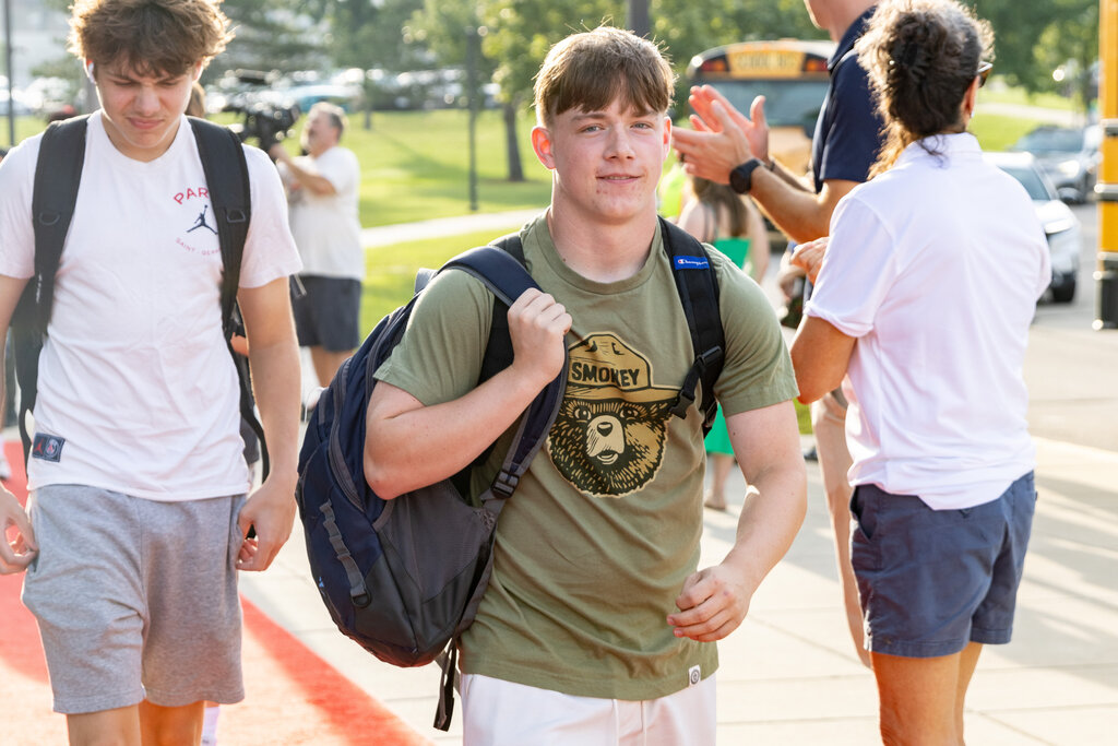 Four individuals walking along a pathway, two carrying backpacks. Red line on ground, background includes school buses and greenery.