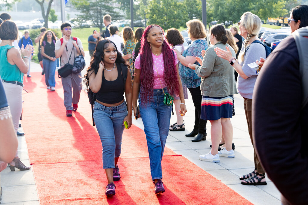 Two women walk down a red carpet outdoors as a crowd gathers behind them. The carpet leads to a building.