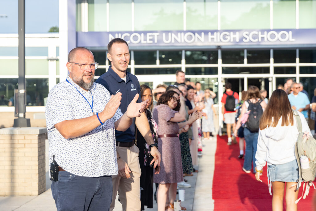 People walk on a red carpet in front of Nicolet Union High School, with a building and glass walls in the background.