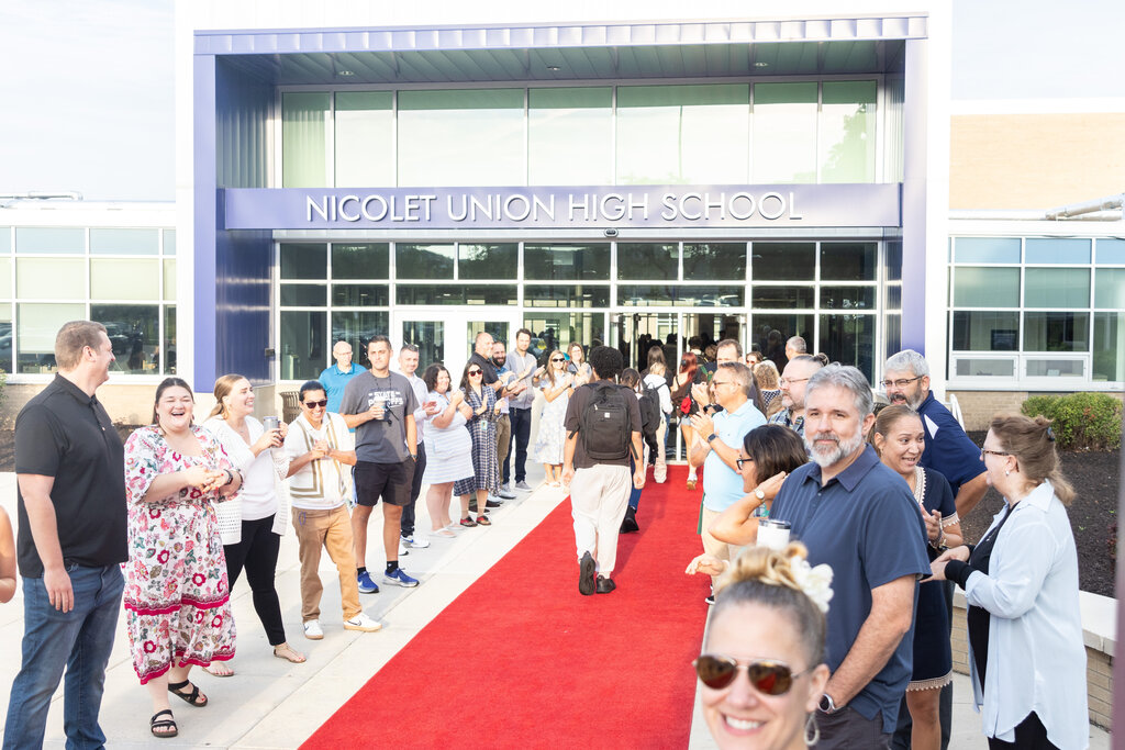 A group of people stand on a red carpet in front of Nicolet Union High School. Some wear glasses, sneakers, and sandals.