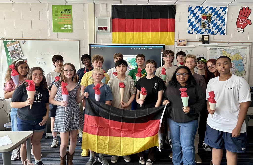 A group of students stands in a classroom, holding a German flag and cups with red lids.
