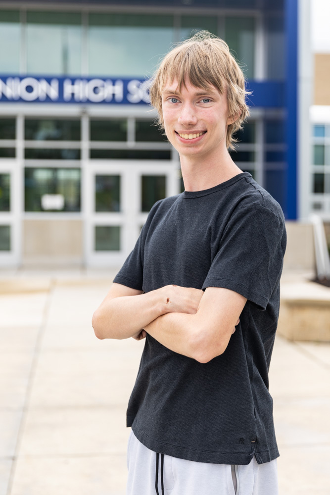 A young man with blond hair smiles outside a high school, wearing a black shirt and white pants.