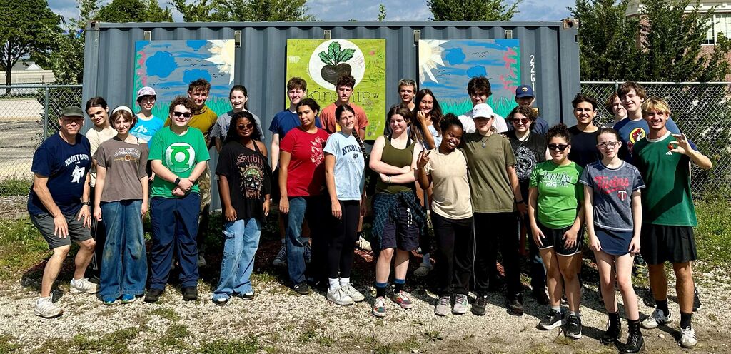 A group of people in casual clothing stands before a large container with murals. Some wear sunglasses.