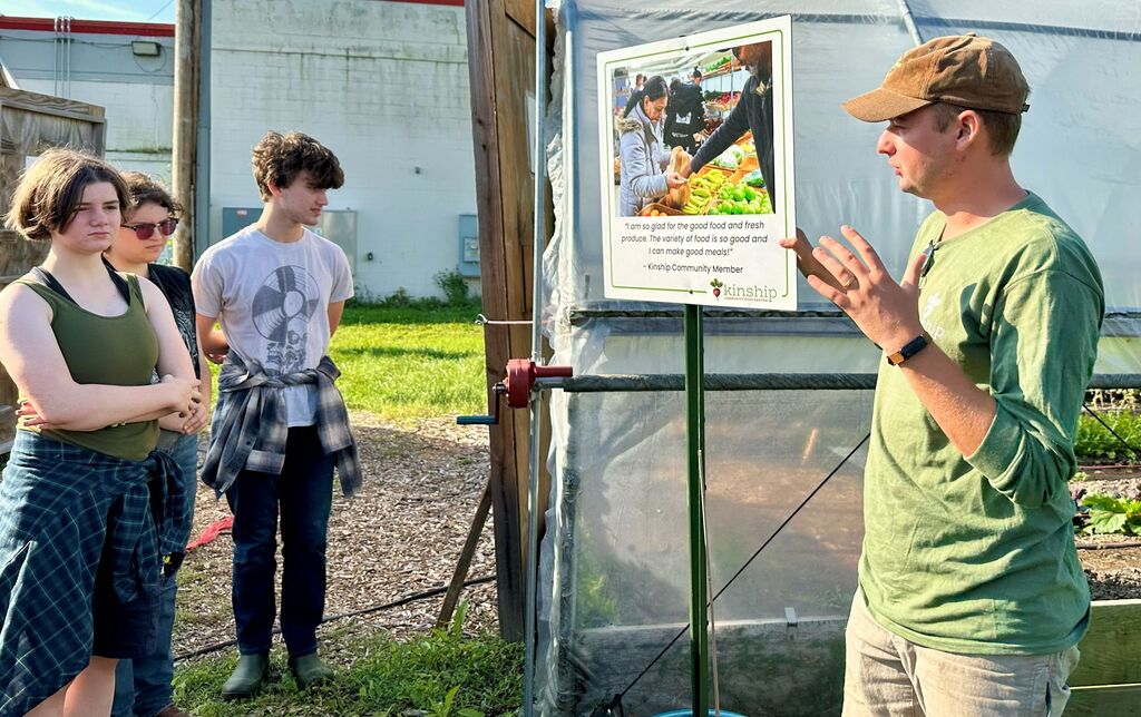 People stand near a greenhouse, one man speaks with a poster on the wall. Grass and plants surround the area.
