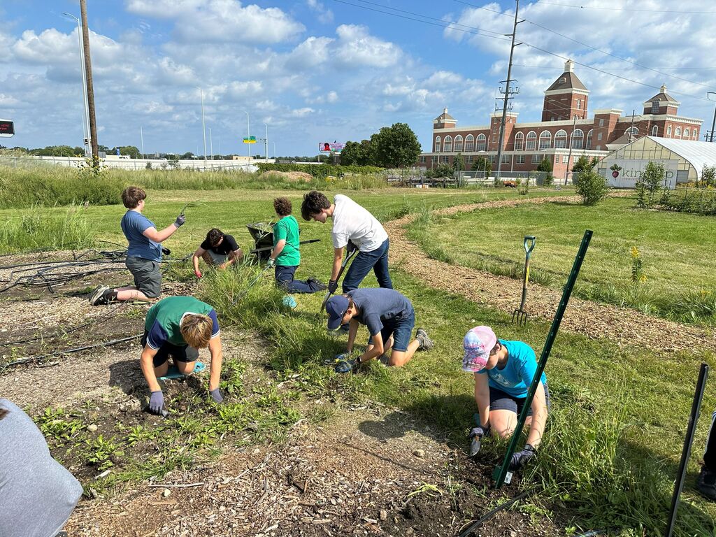 People work in a garden under a partly cloudy sky. Some use shovels. An old building is in the background.