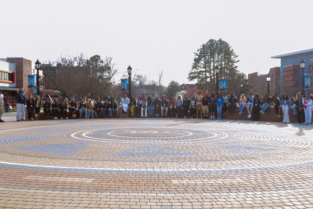 Many people stand around a large circular brick pattern, with trees and buildings in the background.