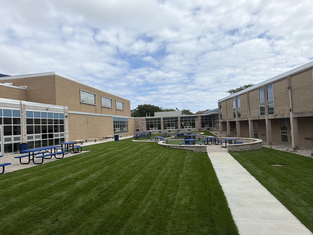 An outdoor area with a grassy lawn and a walkway. Buildings with large windows and a fountain are in the background.