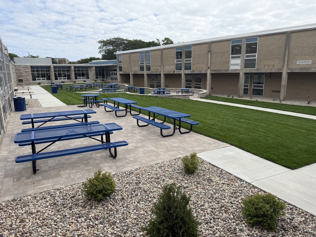 An outdoor area with picnic tables and benches on gravel. A grassy lawn is nearby. A building is in the background.