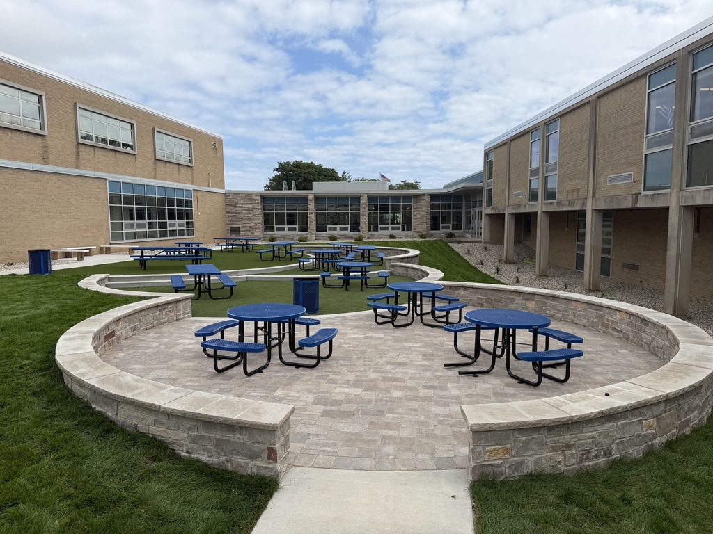 A circular seating area with blue tables and benches on a grassy lawn, framed by a stone border. Buildings with glass windows in the background.
