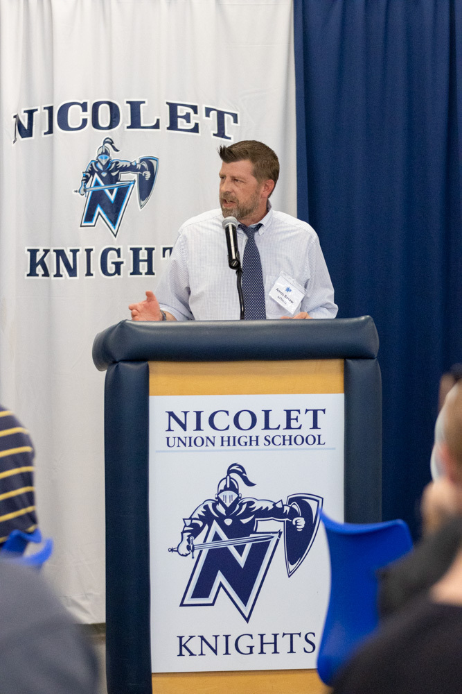 A man speaks into a microphone at a podium with the Nicolet Knights logo. Behind him is a blue and white backdrop.