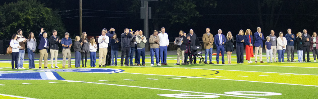 A group of people stands on a football field with illuminated lights in the distance.
